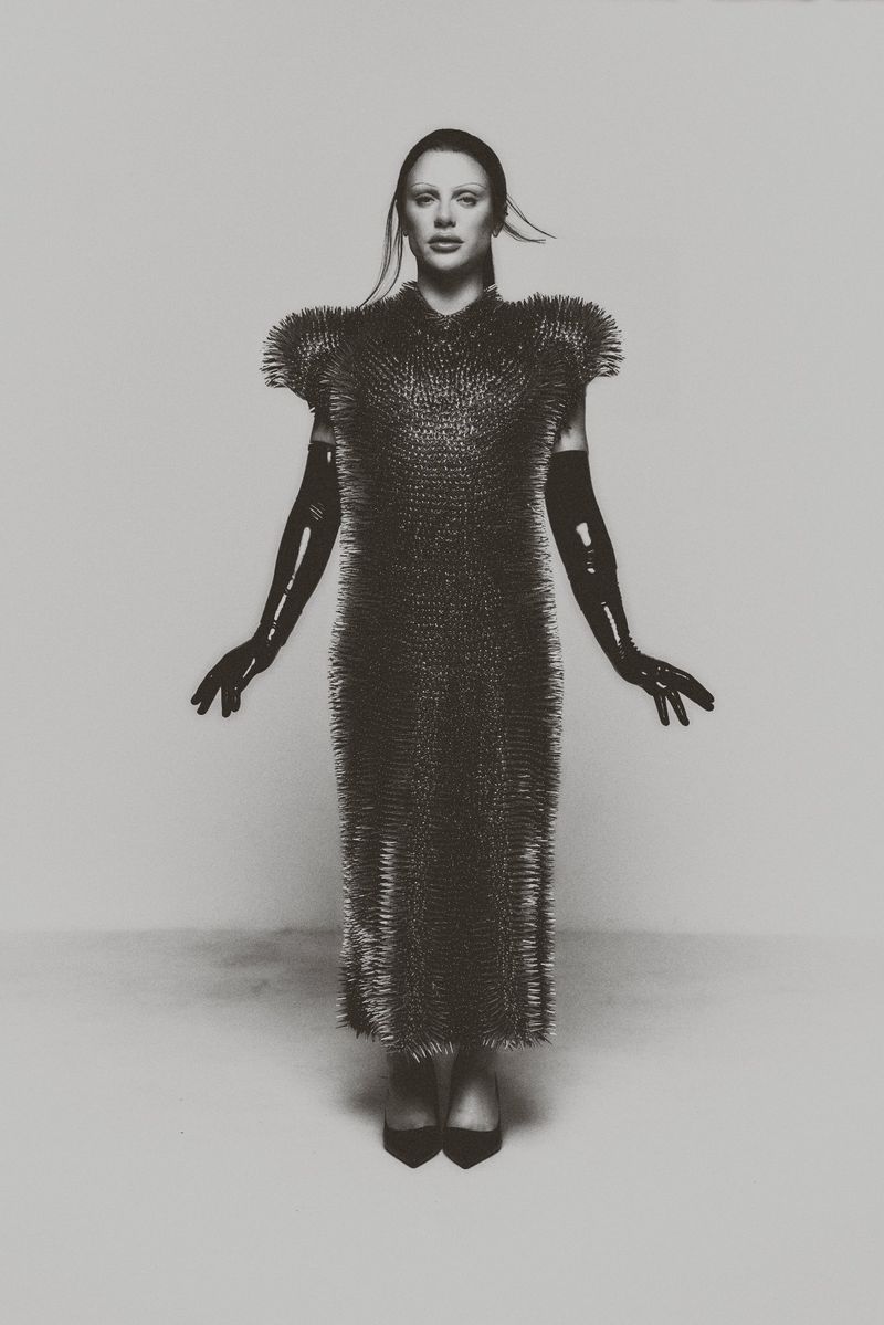 Woman in a floor-length dress covered in spikes, wearing long black gloves and high heels, standing in a photo studio with a light grey background.