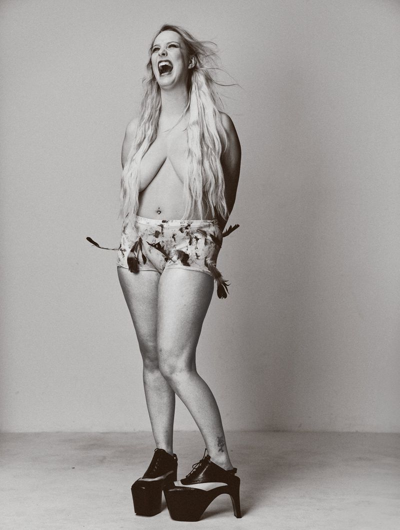 Full body shot of a topless adult woman with blonde hair, wearing high heels and shorts decorated with feathers in a studio setting.