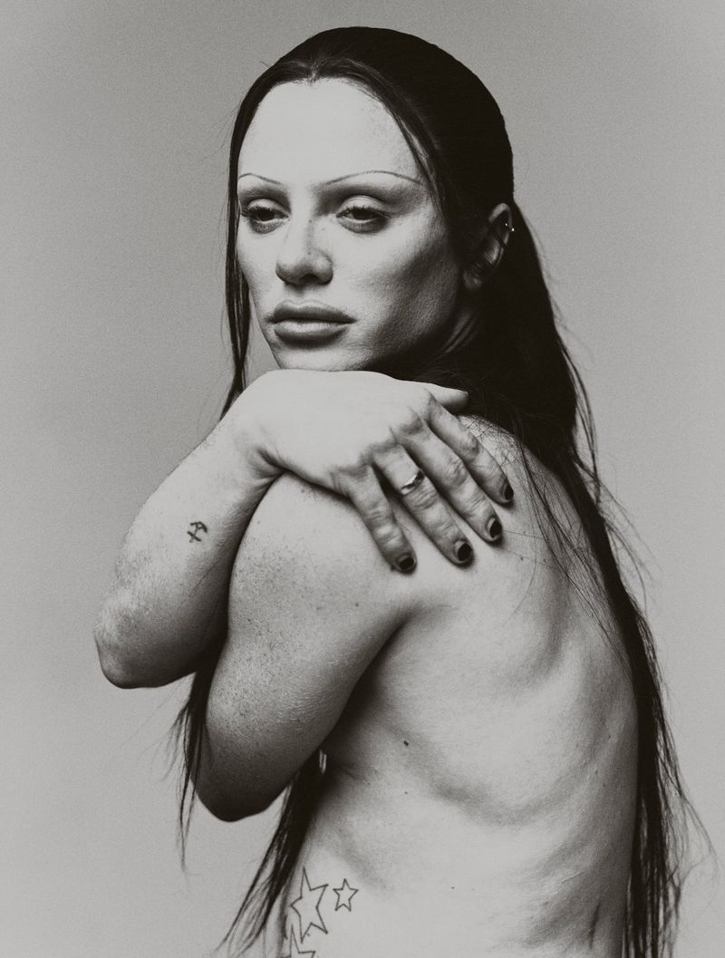 Black and white studio portrait of Bimini with long dark hair, holding their shoulder against a neutral backdrop.