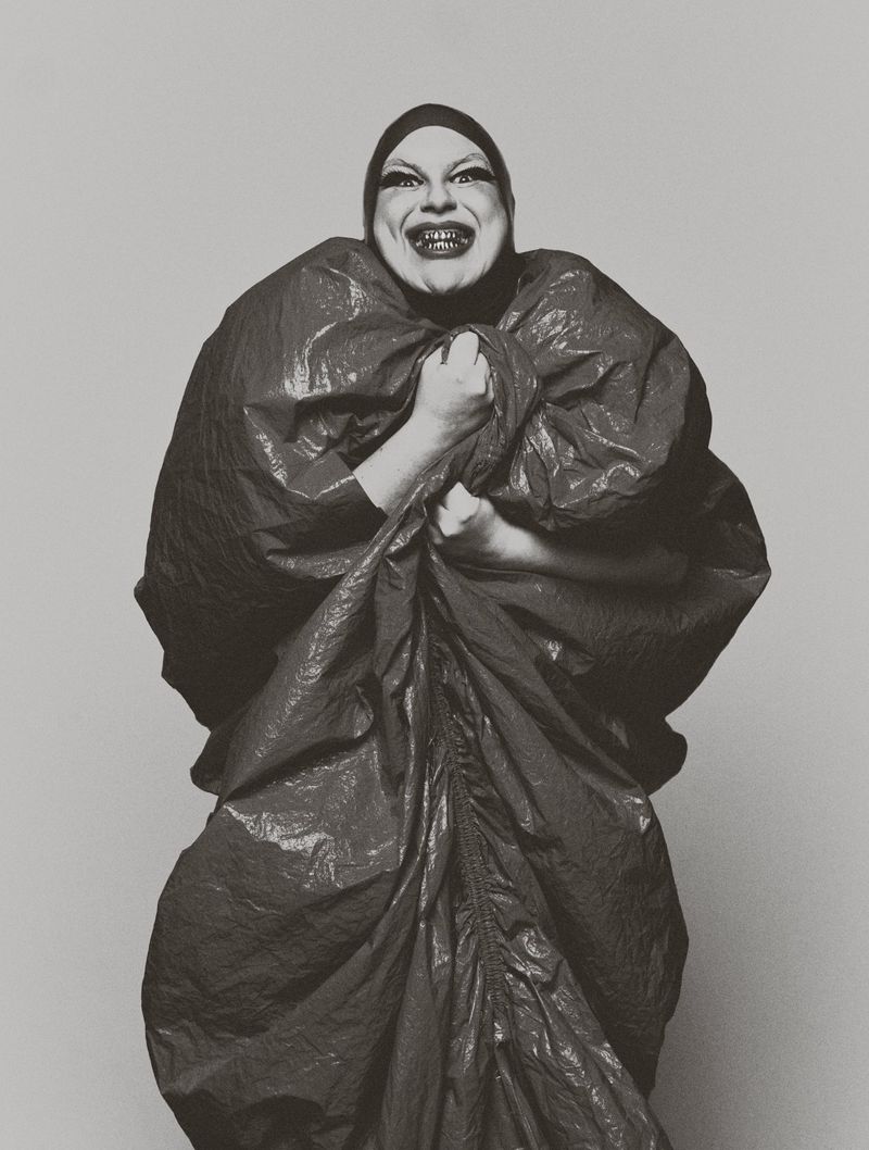 Monochrome portrait of a person with heavy makeup wearing a black head covering and posing in a large draped plastic bag.