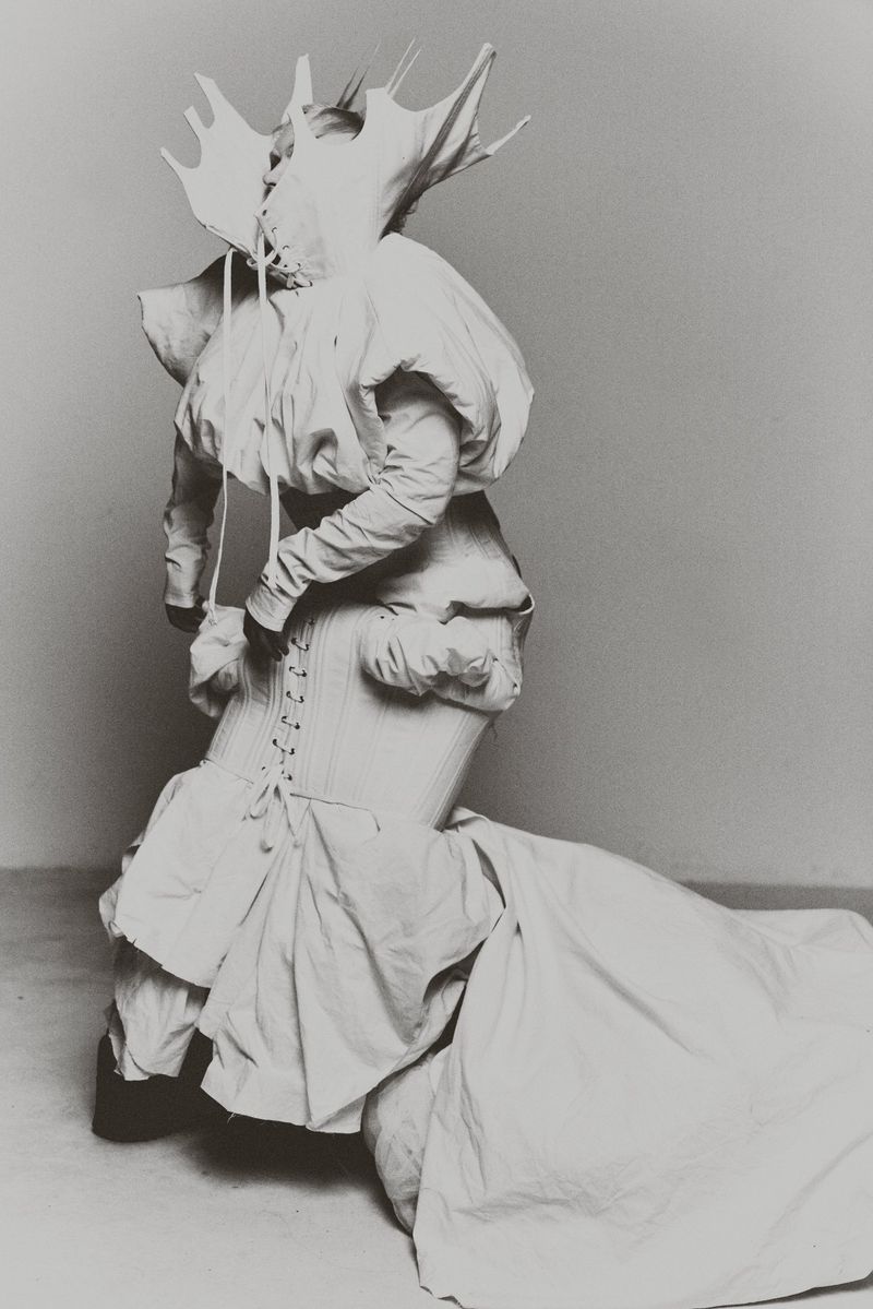 Monochromatic fashion shot featuring a person in a sculptural white garment with an avant-garde headpiece against a grey backdrop.