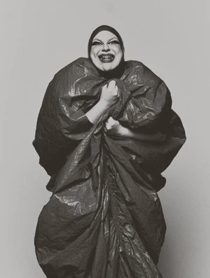 Bimini smiling in an oversized voluminous crinkled coat and tight dark hood against a neutral studio backdrop.