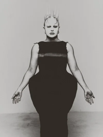Model wearing a structured dark dress with spiked crown hair posing against a neutral backdrop
