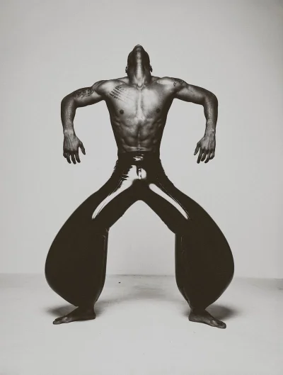 Shirtless man with head tilted back wearing voluminous shiny black trousers against a neutral studio wall.