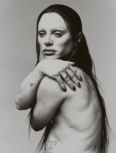 Black and white studio portrait of Bimini with long dark hair, holding their shoulder against a neutral backdrop.