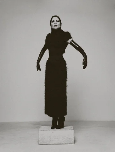 Model Bimini poses on a wooden block in a textured black dress with long black opera gloves against a grey studio background.