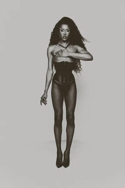 Full-length monochrome studio portrait of Tayce posing in a black corset, fishnet tights, and high heels against a plain background.