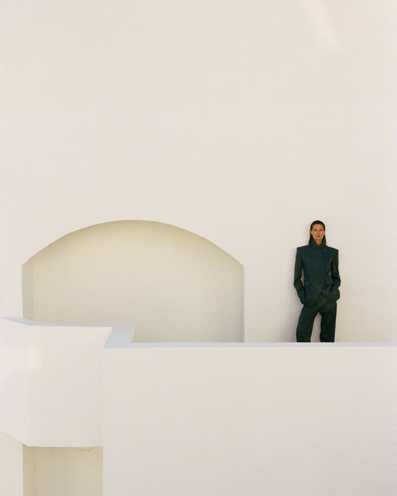 Model Malgosia Bela standing in a dark tailored suit against a minimalist white architectural wall with an arched opening.