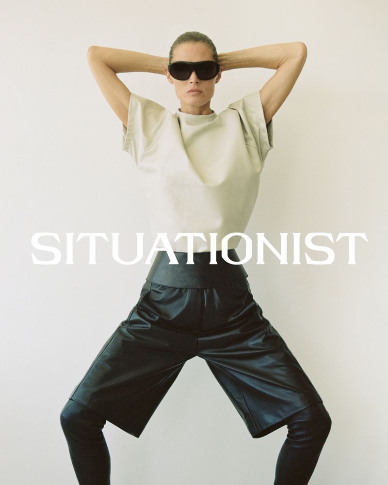 Model wearing dark sunglasses, light short-sleeve top, and black leather shorts against white seamless background