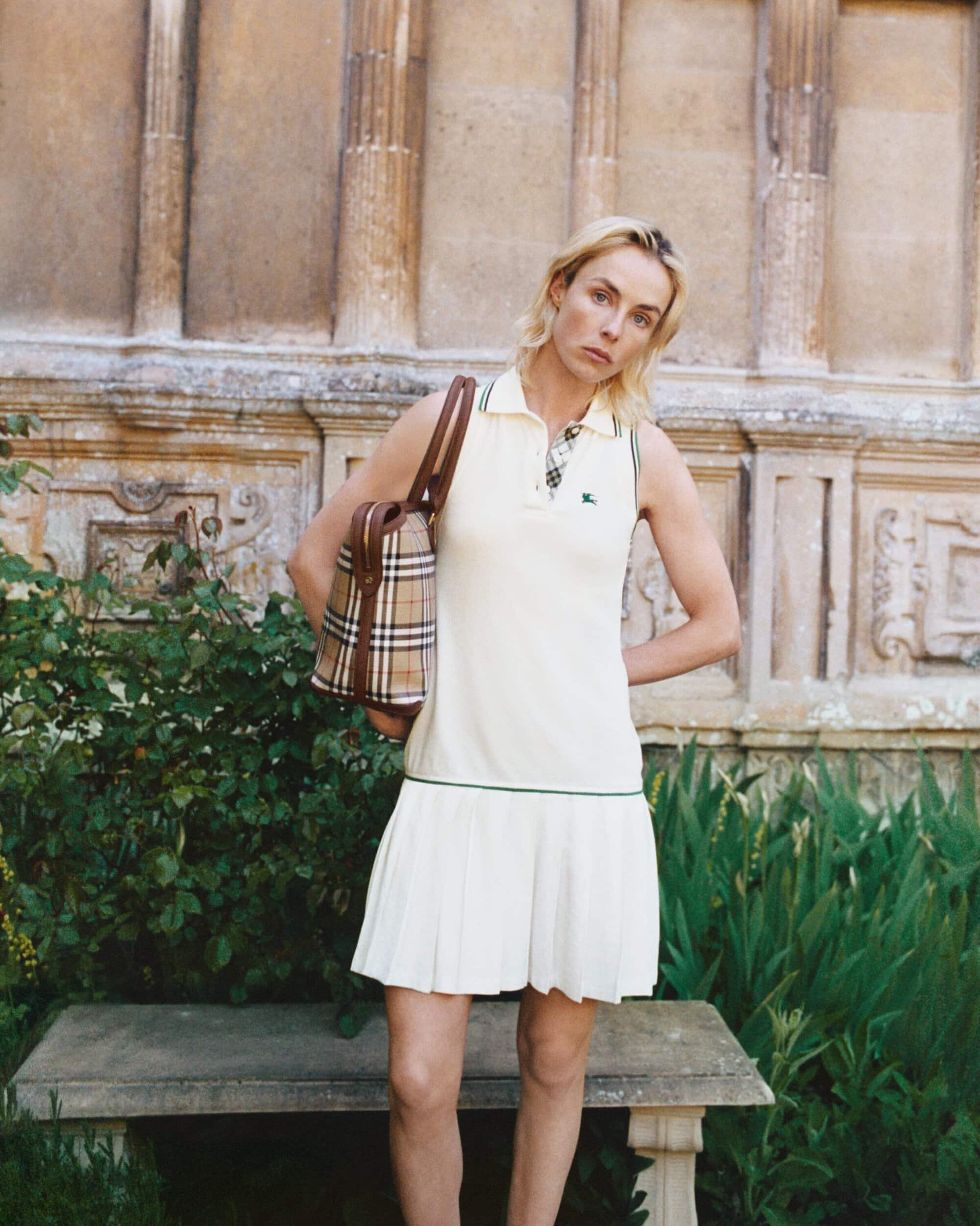 Blonde woman in a white polo dress holding a plaid handbag standing before stone architectural columns and garden foliage