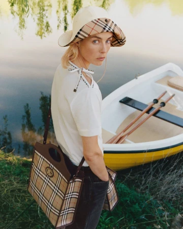 Model Edie Campbell wearing a Burberry check sun hat and carrying a plaid tote bag near a rowboat on a lake shore.