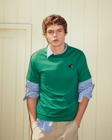 Moses Martin wearing a green t-shirt layered over a blue striped shirt and khaki pants against a pale wood wall.