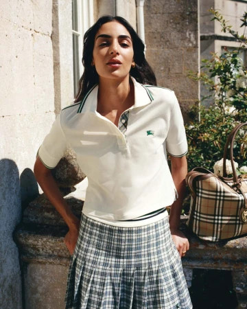Model Nora Attal in a white polo shirt and plaid skirt standing outside a historic stone building with a plaid handbag