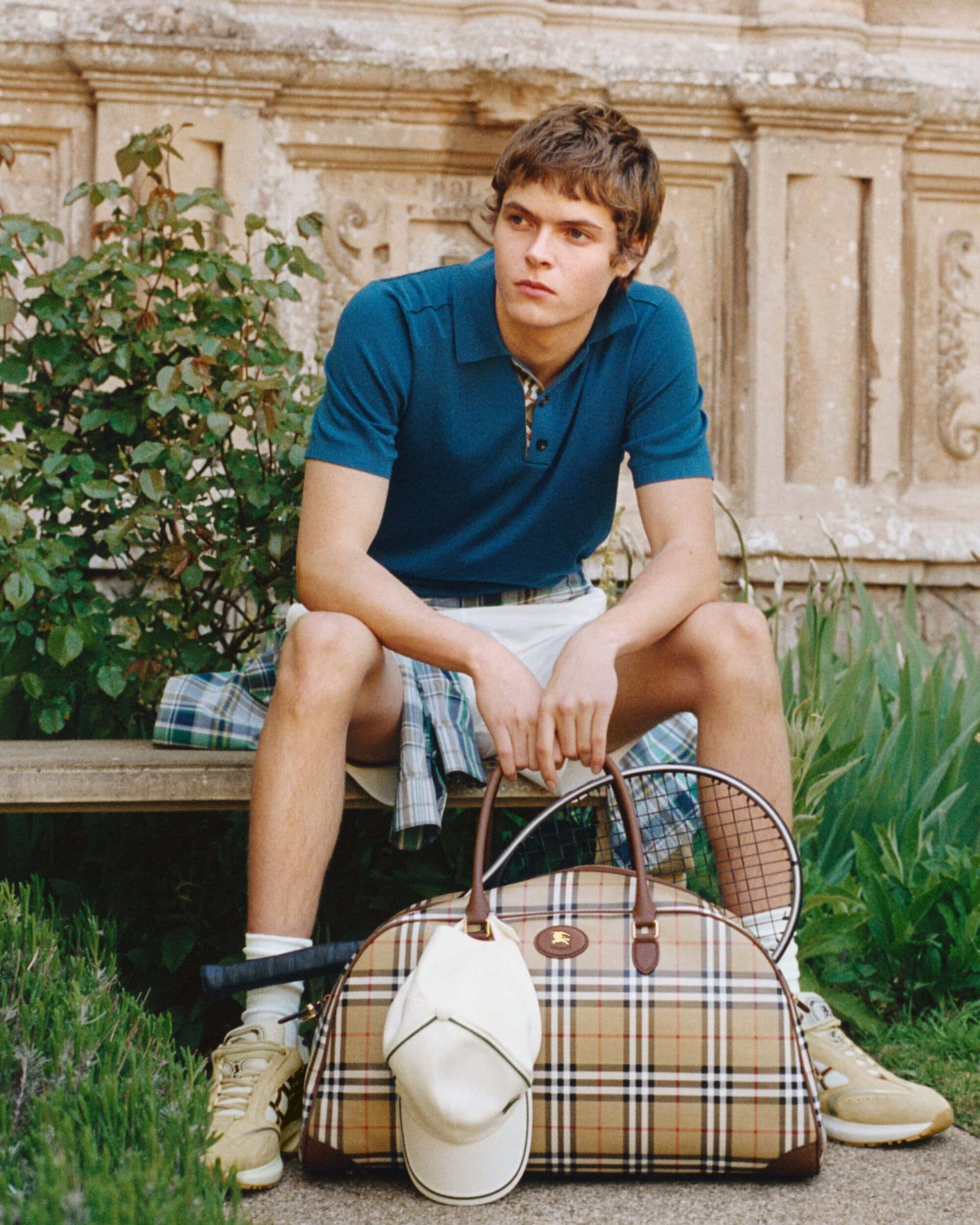 Man in a blue polo shirt and white shorts sits outdoors on a bench beside a plaid check duffle bag and a tennis racket.
