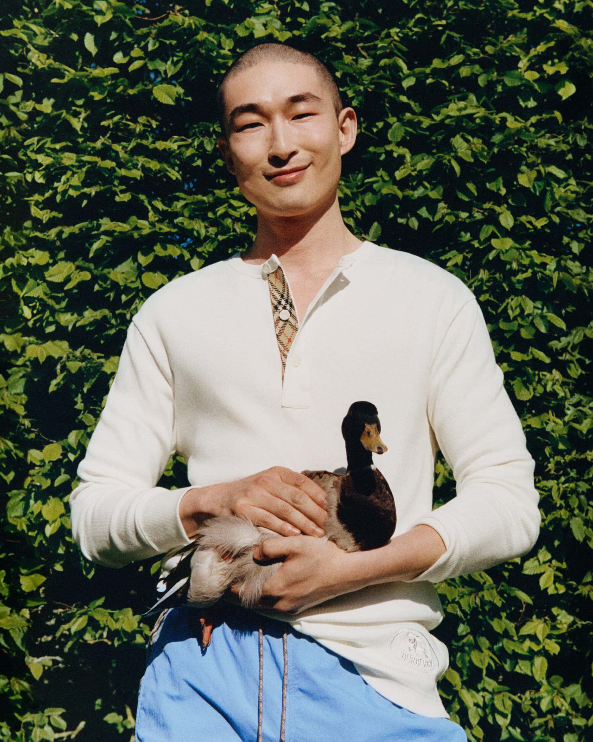 Sang Woo Kim holding a duck while wearing an off-white waffle henley shirt and blue shorts in front of lush green foliage.