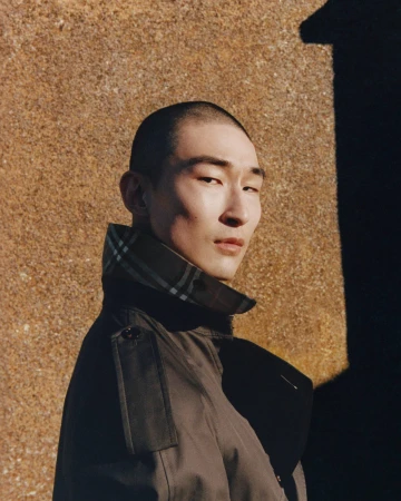 Model Sang Woo Kim wearing a high-collared trench coat with check lining against a textured stone wall in direct sunlight.