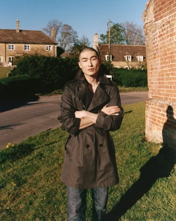 Model Sang Woo Kim wearing a dark double-breasted trench coat standing on a grassy roadside in a countryside setting.