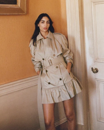 Model Nora Attal leaning against a wall wearing a beige belted mini trench dress with a ruffled hem.