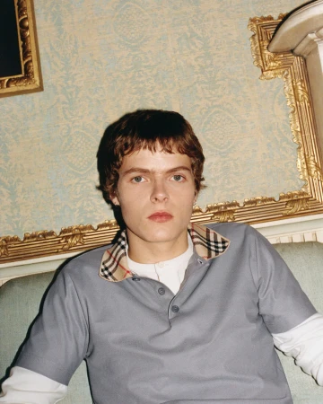 Moses Martin seated wearing a grey Burberry polo shirt with check collar against ornate patterned wallpaper.