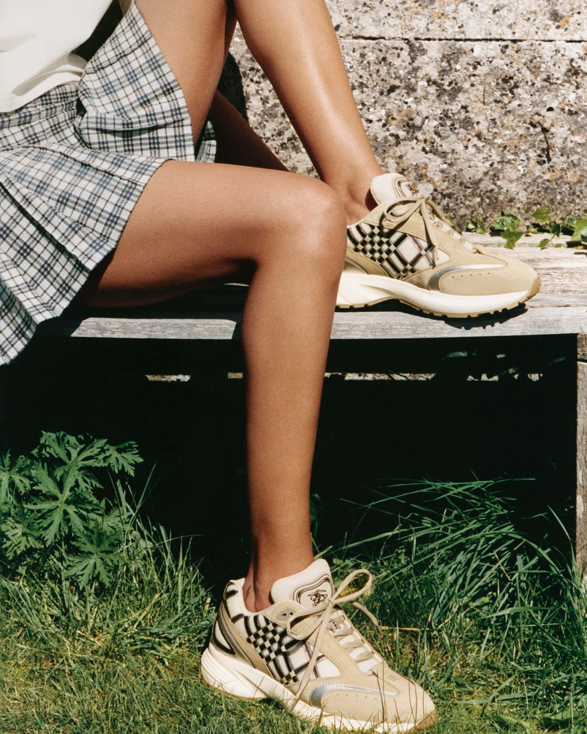 Cropped view of legs wearing beige plaid sneakers and a pleated check skirt, seated on a wooden bench outdoors.