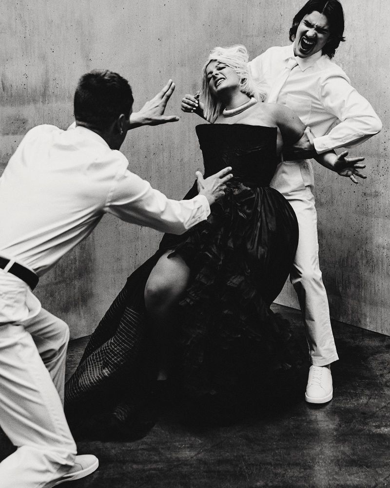 A blond woman in a black strapless gown being held by two men in white shirts against a concrete background.