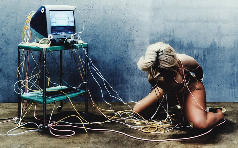 Blonde woman kneeling on the floor wrapped in tangled cables connected to a vintage computer on a rolling cart.