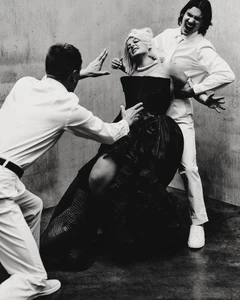 A blond woman in a black strapless gown being held by two men in white shirts against a concrete background.