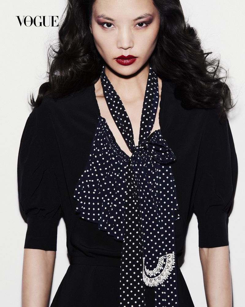 Model Xue Huizi looking directly at the camera wearing a black top and a polka dot necktie against a white backdrop.