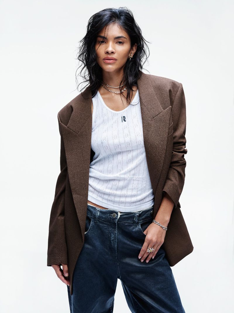 A woman poses in a studio wearing a brown blazer, ribbed tank top, and blue velvet pants against a bright white backdrop.