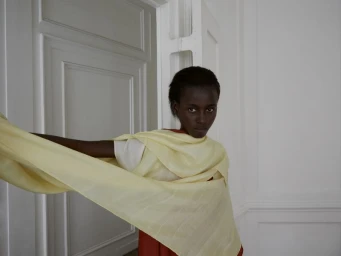 Model Blessing Ifeoluwa poses holding a draped pale yellow fabric wrap in a white architectural interior.