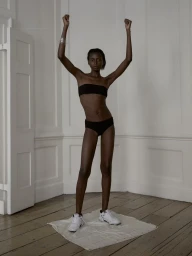 Model Blessing Ifeoluwa standing with arms raised wearing a black bandeau top, black briefs, and white sneakers on a wooden floor.