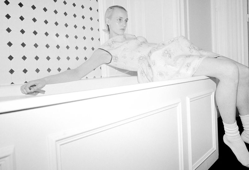 Model George Anderson reclining across the rim of a white bathtub wearing a pale patterned dress and white socks.
