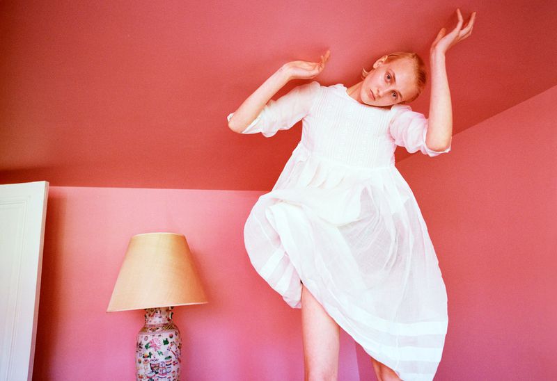 Model George Anderson in a sheer white pleated dress posing under a low pink ceiling beside a porcelain table lamp.