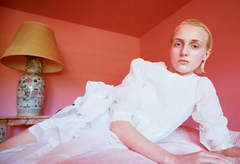 Model George Anderson wearing a white ruffled puff-sleeve dress reclining on a pink bed beside a porcelain table lamp.
