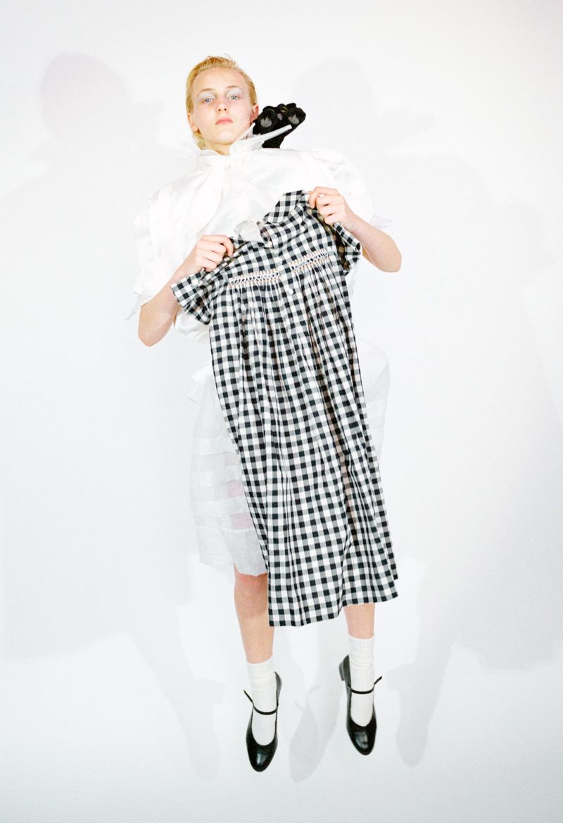 Model holding a black and white gingham check dress in front of a white outfit and black shoes against a white backdrop.