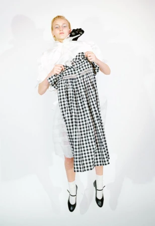Model holding a black and white gingham check dress in front of a white outfit and black shoes against a white backdrop.