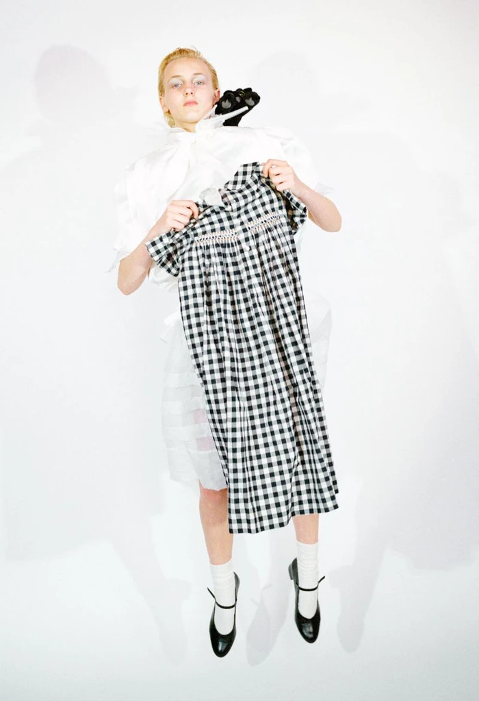 Model holding a black and white gingham check dress in front of a white outfit and black shoes against a white backdrop.