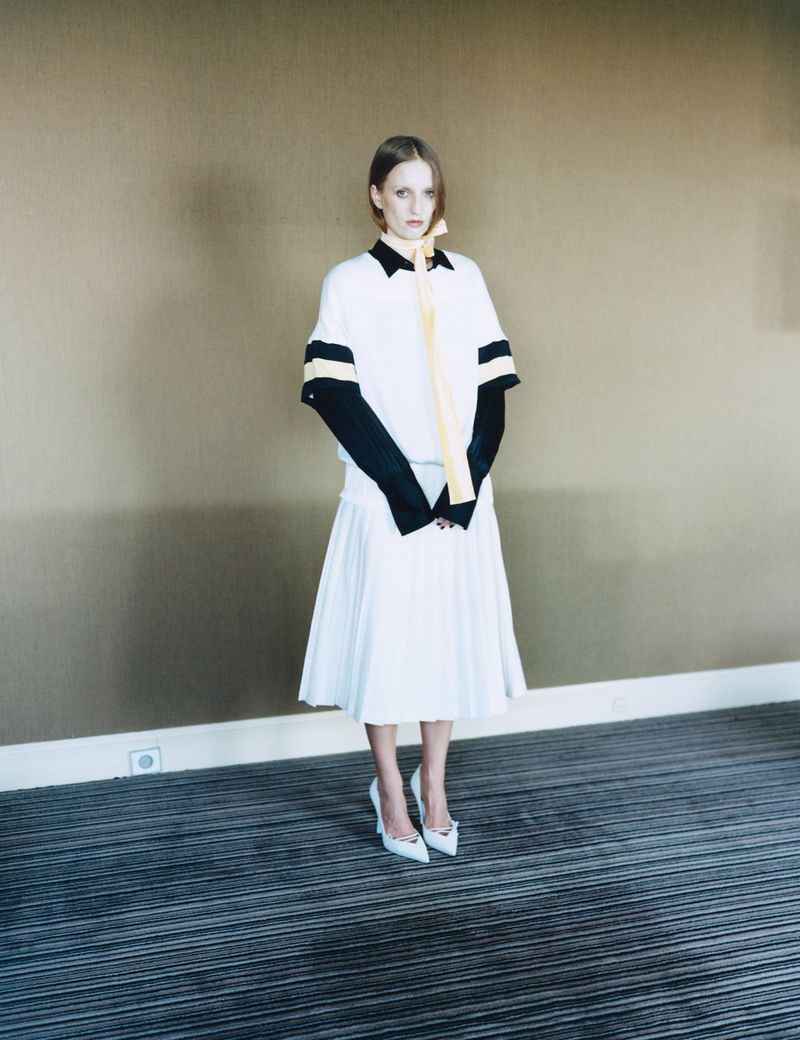 Petra Collins wearing a layered white and black dress with a yellow necktie and white heels against a neutral wall