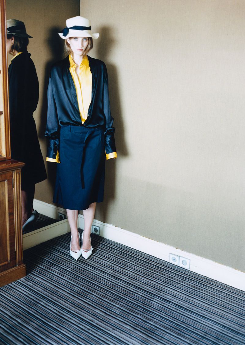 Petra Collins wearing a white hat, black satin shirt, yellow blouse, and navy skirt beside a tall mirror.