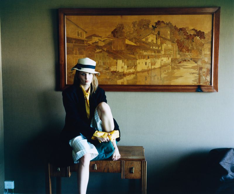 Petra Collins wearing a straw hat, yellow shirt, and black jacket, seated on a wooden console beneath a large framed landscape artwork.