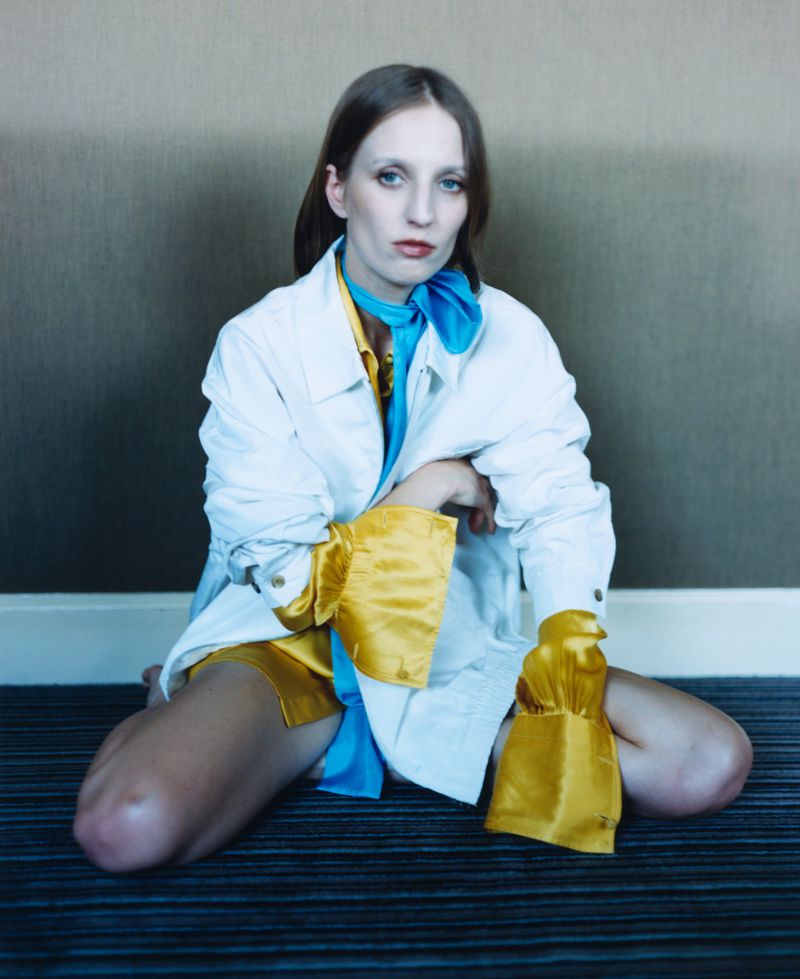 Petra Collins sitting on a striped carpet wearing a white jacket, bright yellow satin cuffs, and a vivid blue scarf tied at the neck.