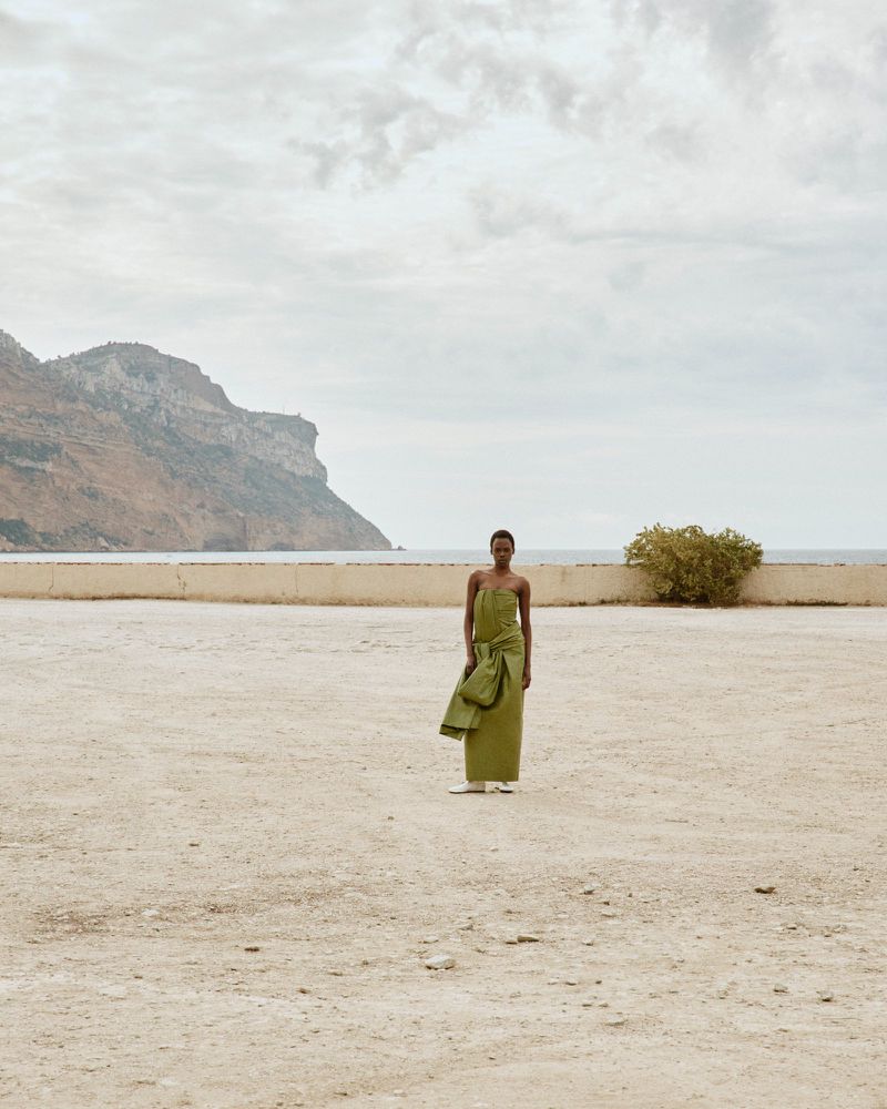 Model Agel Akol in a strapless olive green dress standing on a gravel expanse against a coastal mountain and overcast sky.