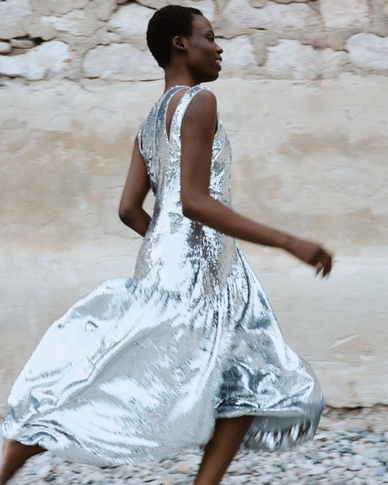Model Agel Akol running in a metallic silver sleeveless dress against a rustic stone wall.