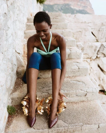 Model Agel Akol seated on stone steps wearing blue leggings, a mint halter top, and shoes with oversized gold ruffled cuffs.