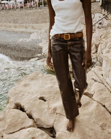 A model walks barefoot on coastal rocks wearing a white tank top, dark trousers, and a belt for Elle Canada.