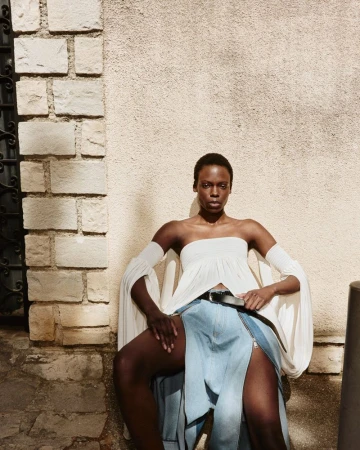 Model Agel Akol seated against a stone wall wearing a white off-the-shoulder draped top and denim skirt for Elle Canada.