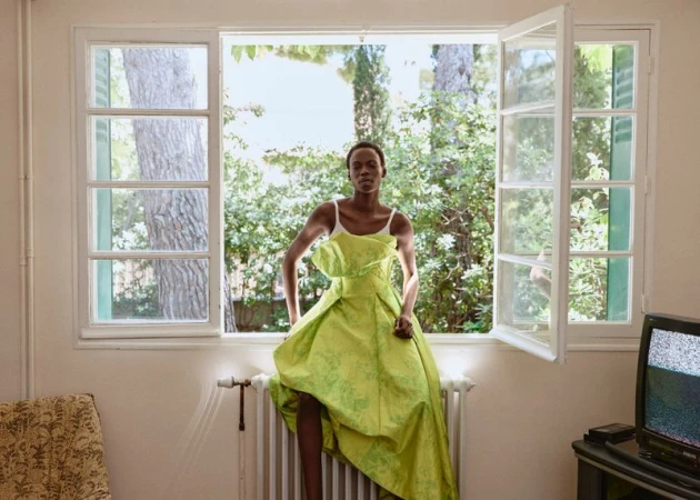 Model Agel Akol sits on a white radiator in front of an open window wearing a voluminous lime green dress for Elle Canada.