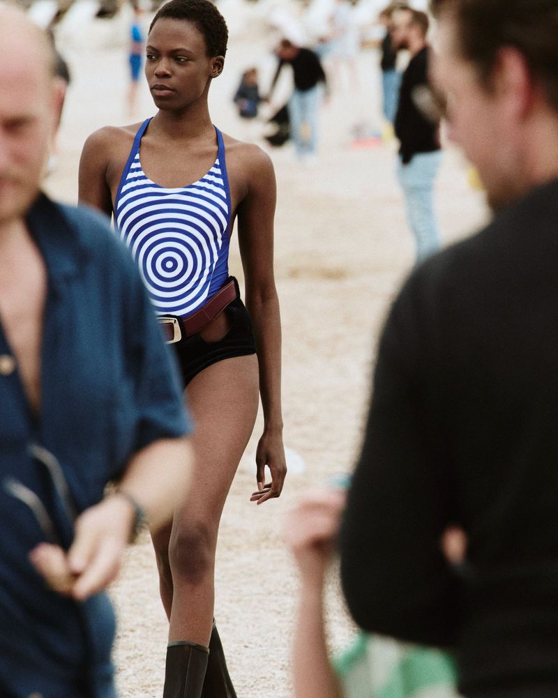 Model Agel Akol walks on a sandy beach wearing a blue and white patterned swimsuit and black shorts for an Elle Canada editorial.