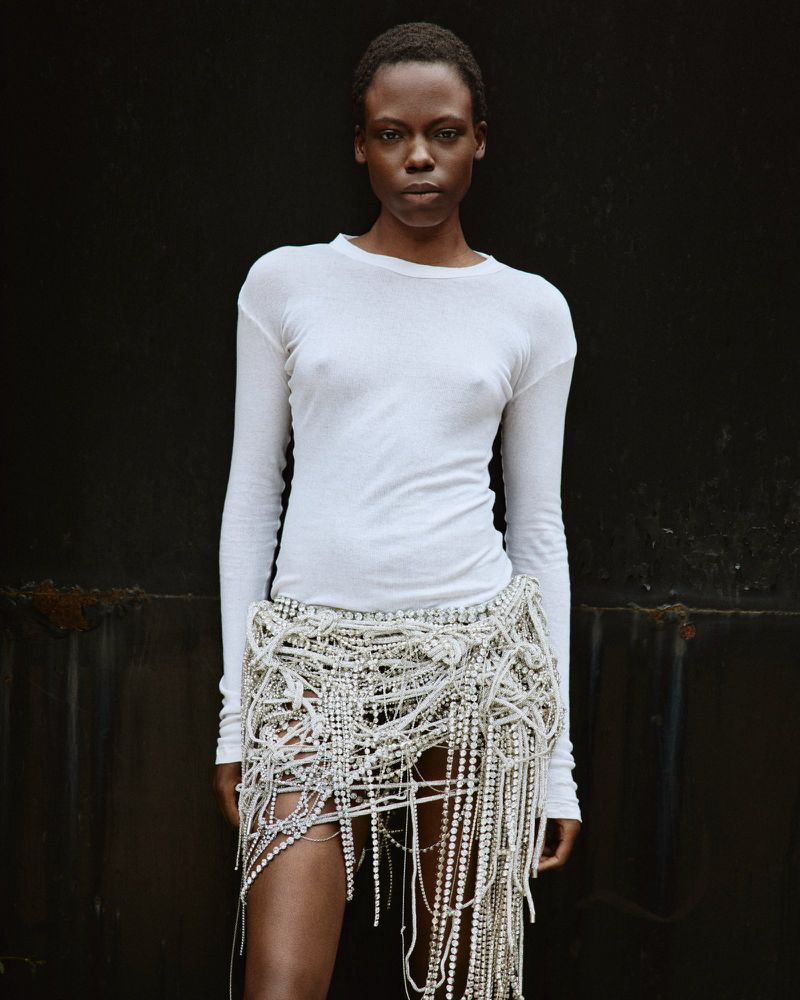 Model Agel Akol in a long-sleeve white top and crystal beaded fringe skirt standing against a dark backdrop.