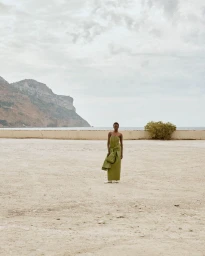 Model Agel Akol in a strapless olive green dress standing on a gravel expanse against a coastal mountain and overcast sky.
